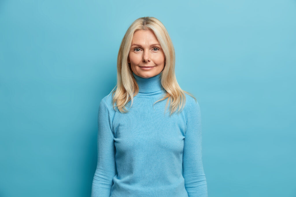 Portrait of beautiful blonde middled aged European woman looks directly at camera dressed in casual poloneck neck smiles gently poses against blue background. Human face expressions concept.